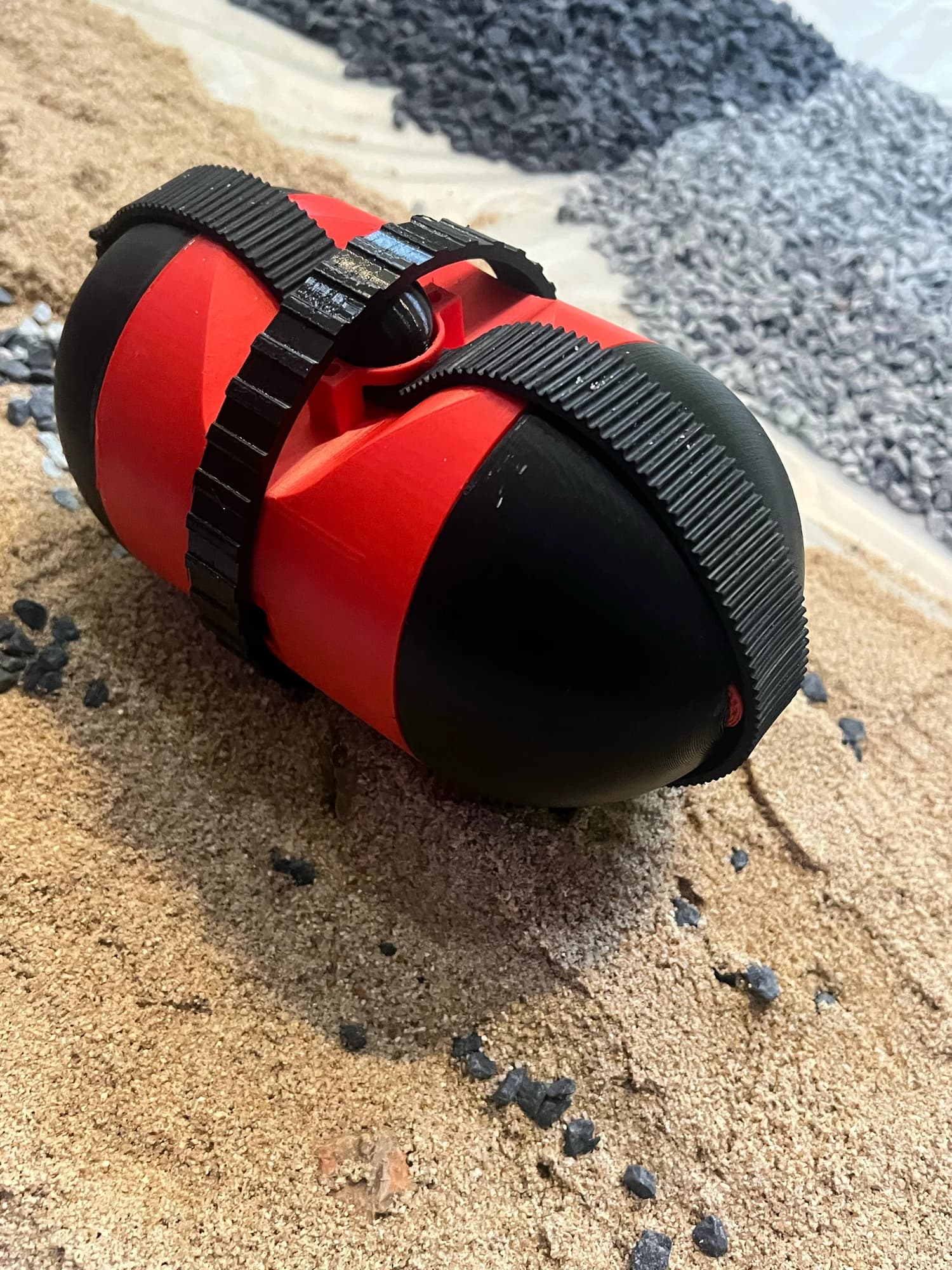 Built red and black capsule robot with perpendicular tracks on a prepared sand-and-gravel surface